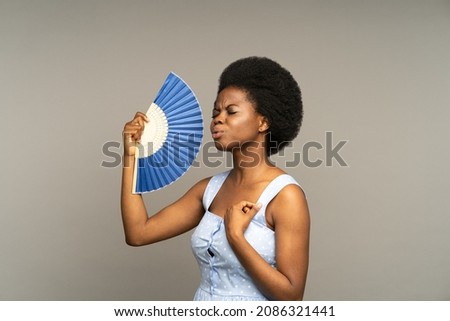 Young afro woman suffering from heat or high air temperature inside, waving with paper fan, standing isolated on grey. Overheated african american female fanning herself to cool down on hot summer day Royalty-Free Stock Photo #2086321441