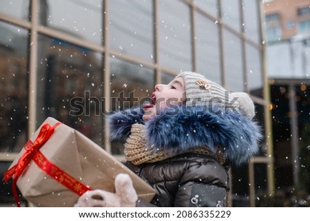 Happy girl with a gift box for Christmas catches snowflakes with his tongue on a city in winter with snow on festive market with decorations and lights. Warm clothes, knitted hat, scarf and fur. 