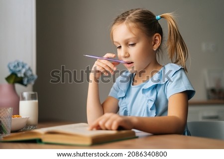 Close-up of thinking pretty elementary child school girl doing homework and holding pen against mouth sitting at home table. Pensive preschool kid reading paper book and writing in copybook. Royalty-Free Stock Photo #2086340800