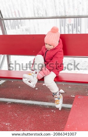 adorable kid girl puts on skates while sitting on a bench near the winter outdoor ice rink