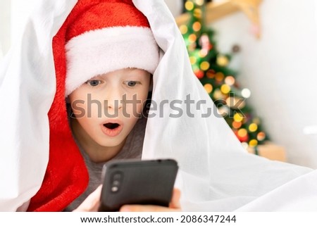 A surprised boy with wide open eyes and mouth lies in bed and looks at the phone screen at home in front of the Christmas tree. A child with a smartphone in his hands saw the shocking good news.