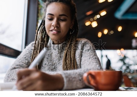 Creative female poet develop own talent while writing text ideas in textbook enjoying time in coffee shop, dark skinned hipster girl with braiding pigtails learning and planning in cafeteria Royalty-Free Stock Photo #2086364572