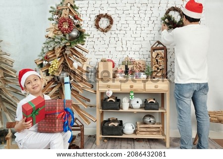 Happy boy putting presents under Christmas tree when his father hanging wreath on wall in background