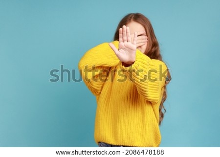 Little girl covers eyes with hand, shows stop gesture,feels shame or fear to watch forbidden content, wearing yellow casual style sweater. Indoor studio shot isolated on blue background. Royalty-Free Stock Photo #2086478188