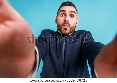Funny bearded businessman wearing official style suit, taking selfie, looking at camera POV, point of view of photo, sending air kiss. Indoor studio shot isolated on blue background.