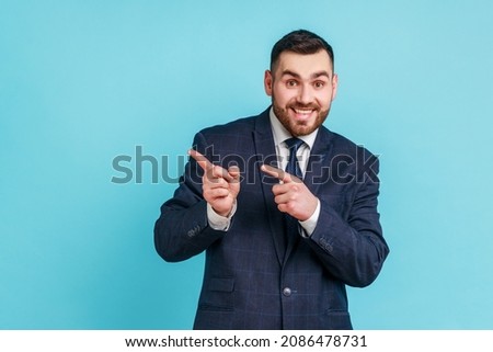 Advertise here! Positive bearded man wearing official style suit pointing finger away paying your attention at empty space for advertisement. Indoor studio shot isolated on blue background. Royalty-Free Stock Photo #2086478731