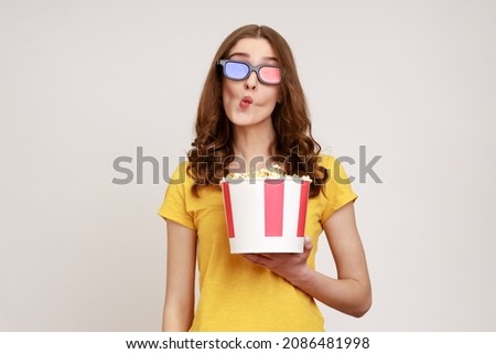 Young childish woman in 3d glasses and yellow casual t-shirt, watching movie film, holding bucket of popcorn, making fish face grimace with pout lips. Indoor studio shot isolated on gray background.