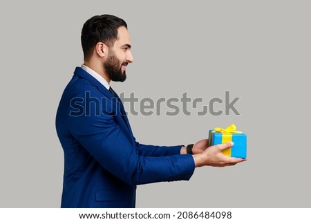 Side view portrait of smiling positive man giving blue wrapped present box, congratulating with holidays, holding gift, wearing official style suit. Indoor studio shot isolated on gray background.