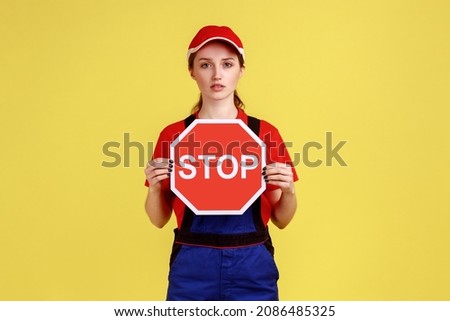 Portrait of serious worker woman standing showing red stop sign, looking at camera with confident strict expression, wearing overalls and red cap. Indoor studio shot isolated on yellow background.