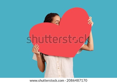 Portrait of unknown woman holding big red heart in hands, peeping from love symbol, being shy to tell about her feelings, wearing white shirt. Indoor studio shot isolated on blue background.