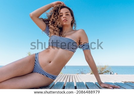 beautiful young hispanic latin girl, in swimsuit sitting looking at the camera, enjoying a sunny day at the beach, with the sea behind her, travel and tourism concept.