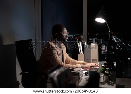 Busy young African man typing on computer keyboard and looking at screen while working late in office Royalty-Free Stock Photo #2086490143