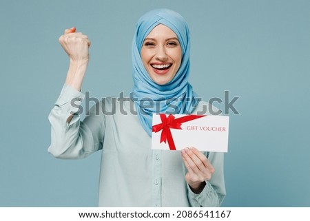 Young arabian asian muslim woman in abaya hijab hold store gift certificate coupon voucher card do winner gesture isolated on plain blue background. People uae middle eastern islam religious concept