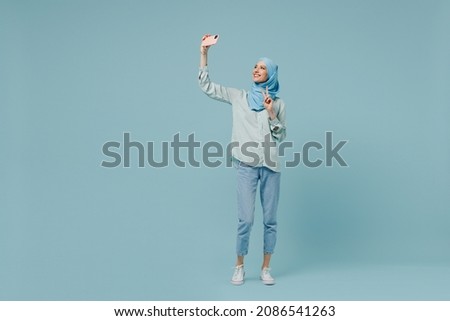 Full body fun young arabian asian muslim woman in abaya hijab doing selfie shot on mobile cell phone show v-sign isolated on plain blue background. People uae middle eastern islam religious concept