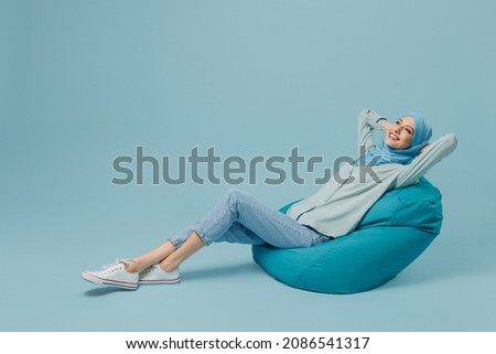 Full body relaxed happy cheerful young arabian asian muslim woman in abaya hijab sit in bag chair isolated on plain blue background studio portrait. People uae middle eastern islam religious concept Royalty-Free Stock Photo #2086541317