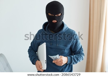 Indoor shot of burglar robbed the house and leaves with the stolen laptop and money, male burglar wearing dark blue hoodie and black mask, performed illegal actions.