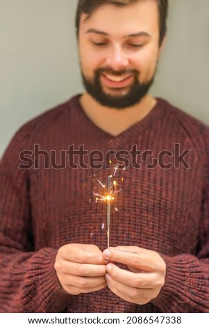 Handsome young man with a beard celebrates the new year in a cozy burgundy marsal knitted sweater and holds a sparkler in his hands smiling in bokeh. Concept: celebrating Merry Christmas and New Year