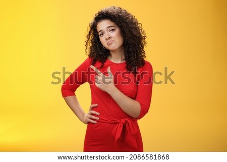 Upset disappointed and unimpressed attractive young european curly-haired woman in red dress pursing lips in gloomy face, frowning displeased as pointing at upper left corner from regret and dislike Royalty-Free Stock Photo #2086581868
