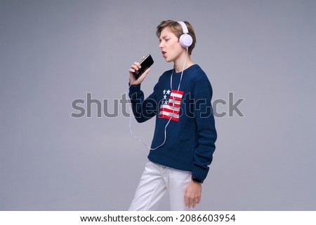 Young expressive singer lover joyful fun caucasian man in casual basic headphones wearing sweater sings voice recording song using mobile cell phone dictaphone isolated on gray studio background.