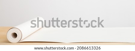 Opened white sheet of wallpaper roll on wooden table at light gray wall background. Closeup. Front view. Preparation for repair work of home. Empty place for text. Wide banner. Royalty-Free Stock Photo #2086613326