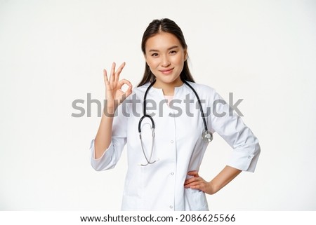 Very good. Smiling confident asian female doctor, showing okay, ok sign in approval, confirm smth, saying yes, give approval, standing in medical uniform, white background