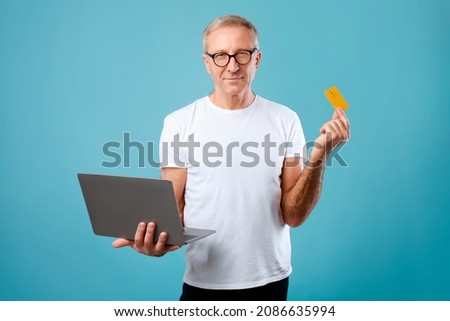 Portrait of confident senior man in eyeglasses holding laptop and showing credit card on blue studio background, adult male using bankcard and personal computer, shopping or banking online, copy space