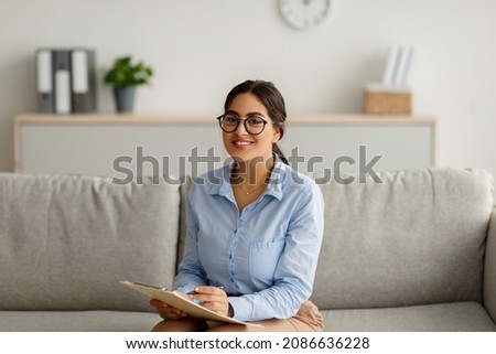 Cheerful arab female psychologist writing in clipboard, having session with client, sitting on couch in office. Friendly psychotherapist taking notes during consultation in mental health clinic