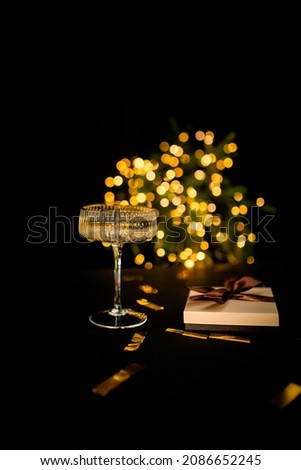 A glass of champagne on a black background with lights, a gift box lies nearby.