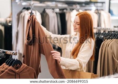 Side view of cheerful pretty young woman buyer choosing clothes from rack in clothing store, blurred background. Cute female shopaholic select and buying clothes in fashion boutique during sale. Royalty-Free Stock Photo #2086699315
