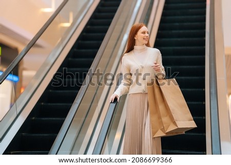 Low-angle view of smiling young woman holding on escalator handrail and riding escalator going up in shopping mall, looking away, paper bags with purchases in hands, blurred background. Royalty-Free Stock Photo #2086699366