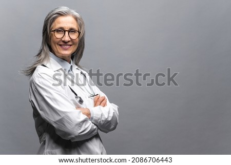Headshot of intelligent professional mature female doctor with gray hair, wearing glasses, medical white gown and stethoscope, looking at the camera, therapist physician standing isolated on gray Royalty-Free Stock Photo #2086706443