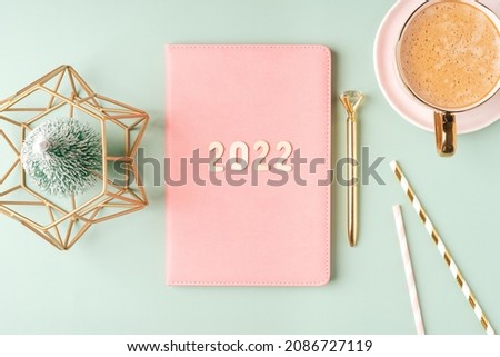 Top view Desktop Christmas pink notepad with 2022 letters text. Flat lay on green mint table background with planner, cup of coffee, candle, Christmas decoration, notebook and stationery.
