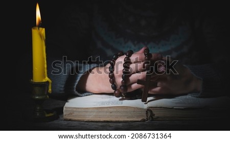 Close up hands of young beautiful girl praying to God with wooden rosary in a candlelight. Selective focus on cross.