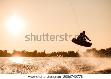 Great view of dark silhouette of active male rider holding rope and making extreme jump on wakeboard over splashing water. Wakeboarding and water sports activity. Royalty-Free Stock Photo #2086740778
