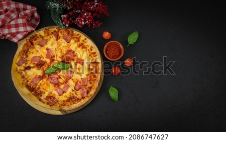 Round baked pizza on a wooden cutting board, red checkered kitchen napkin, ketchup, basil leaves and cherry tomato on the left on a black background with copy space on the right top view close-up.