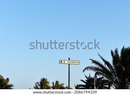 Road sign with airplane icon, direction arrow and word in Spanish "Aeropuerto" (in English: Airport)  fixed on high post amidst tops of green palms in background of sky in Lanzarote, Canary islands