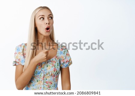 Surprised blond girl, pointing and looking at upper right corner, watching smth amazing, checking out sale advertisement, standing over white background