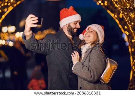 Christmas lovers laugh and take selfies outdoors on Christmas eve. A young happy couple with Santa hats on heads standing on the street and taking selfies on New Year's eve.