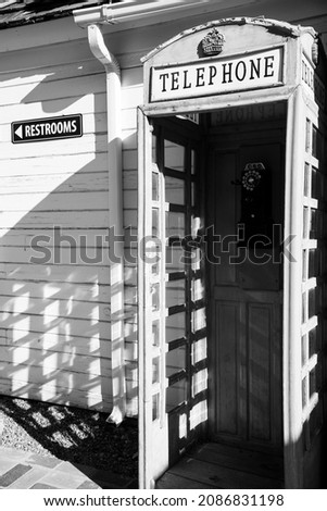  Black and white photo of a vintage telephone booth against a white building.