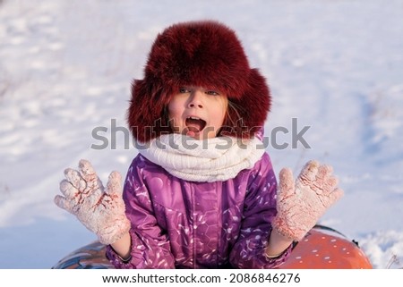 Portrait of a happy girl in a burgundy fur hat, looking into the camera, on a sunny winter day. The girl is enjoying a winter, frosty day. Winter holidays, sledding.