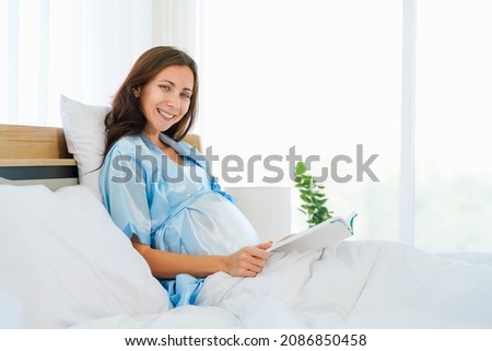 portrait of happy pregnant woman sitting on bed reading book at home in bedroom