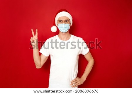 concept - people, protection from covid 19 virus. man in medical mask, Santa Claus xmas hat, shows two fingers with V-shaped peace victory sign with his hand. isolated on red background space for text