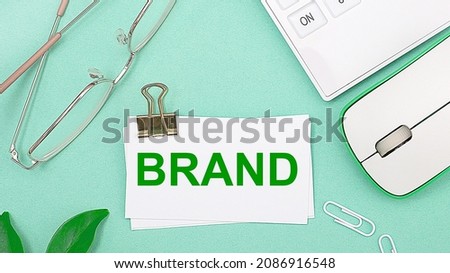 On a light green background there is a white calculator, a computer mouse, green leaves of a plant, gold-rimmed glasses and a white card with text BRAND. Business concept