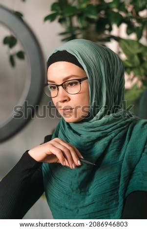 Serious young muslim woman in a hijab leads her video blog, live broadcast.Cozy home interior with indoor plants.Social media and technology concept,diverse people.Selective focus.