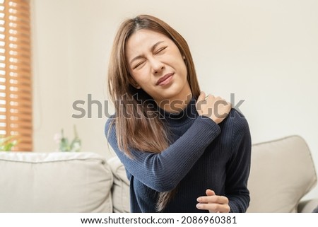 Body muscles stiff problem, asian young attractive woman, girl pain with back pain ache from work, holding massaging rubbing shoulder hurt or sore, painful sitting on sofa at home. Healthcare people. Royalty-Free Stock Photo #2086960381