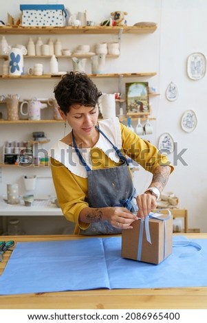 Happy female artist packing gift with handmade pottery after master class in ceramic studio. Young woman entrepreneur packaging for delivery handicraft tableware. Small business and art shop ownership