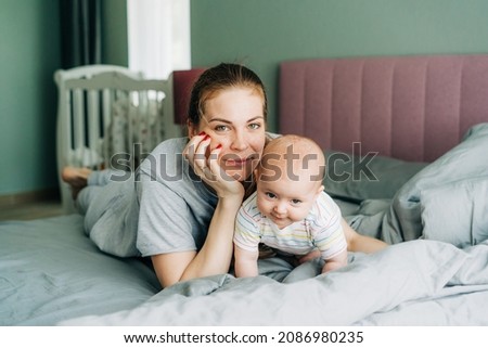 Portrait of a young Caucasian beautiful mom and baby on the bed in the bedroom. Domestic live, mother-child relationship, newborn care, maternal responsibilities.