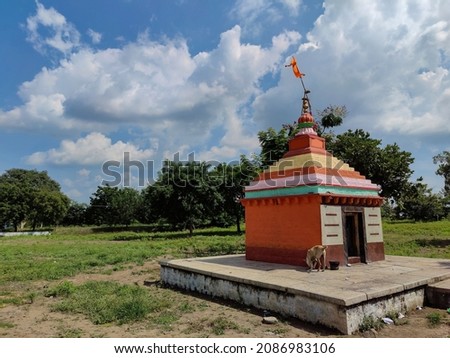 Stock photo small old hindu temple painted with orange color, situated in the middle of grass land, surrounded by green trees, blue sky with white clouds on background. Picture captured at Gulbarga.