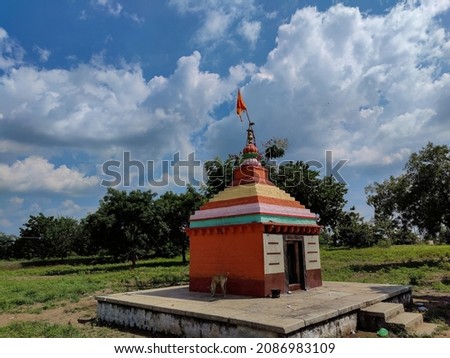 Stock photo small old hindu temple painted with orange color, situated in the middle of grass land, surrounded by green trees, blue sky with white clouds on background. Picture captured at Gulbarga.