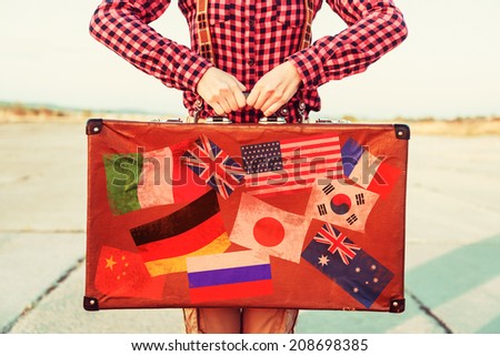 Woman holds brown vintage suitcase, face is not visible. Suitcase with stamps flags representing each country traveled.